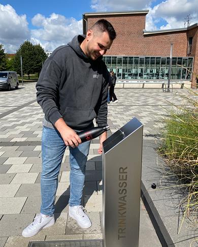 Ein junger Mann steht an einem Trinkbrunnen und füllt eine Trinkflasche auf